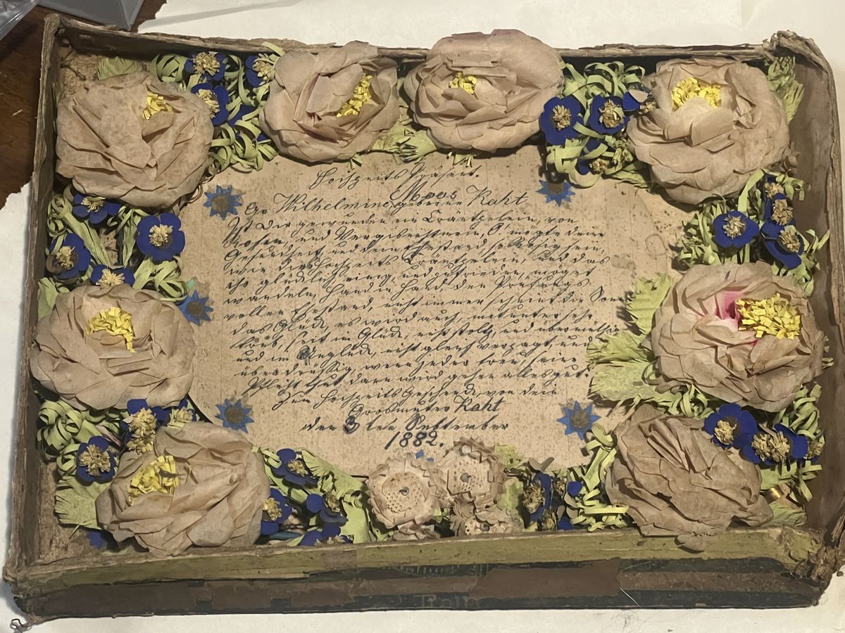 A shallow box holding a handwritten German document surrounded by handmade paper roses and blue paper flowers
