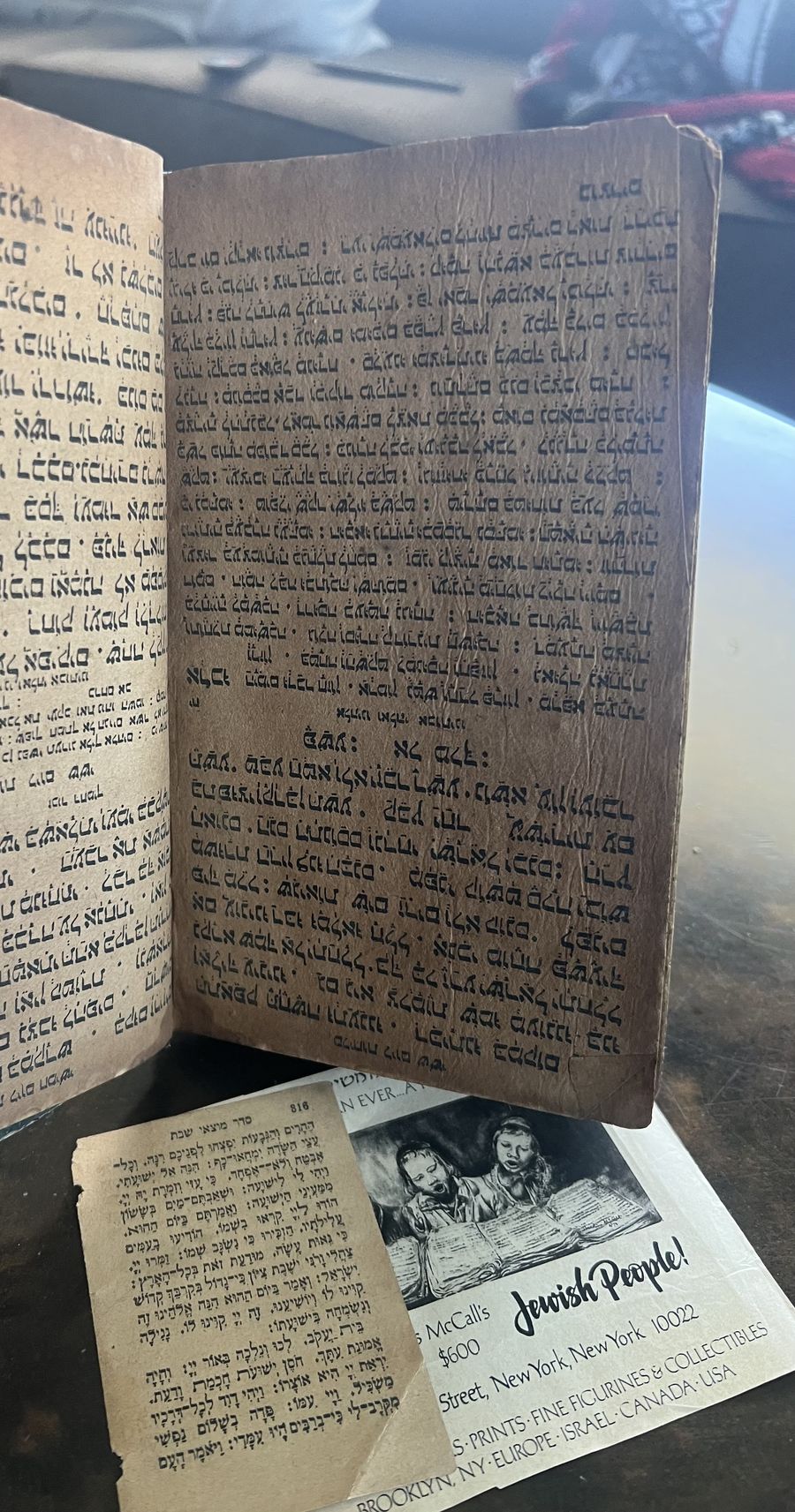 The book propped open above a Hebrew prayer-book leaf and a printed advertisement for a Judaica prints and collectibles dealer