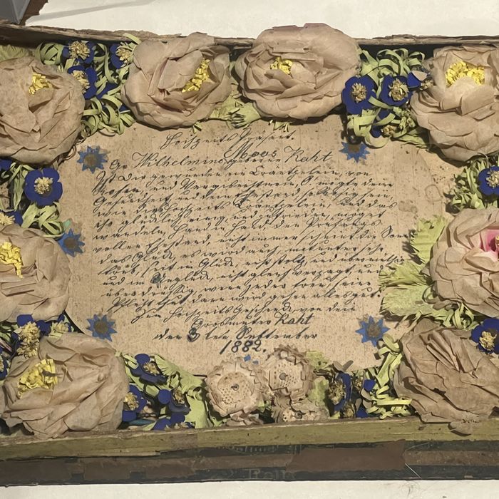 Handmade paper roses and blue paper flowers wreathed around a handwritten German document dated 1882