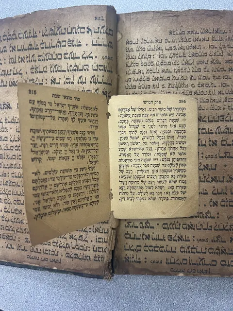 Two loose printed leaves in square Hebrew type with vowel points lying on the open pages of the hand-written Syriac book
