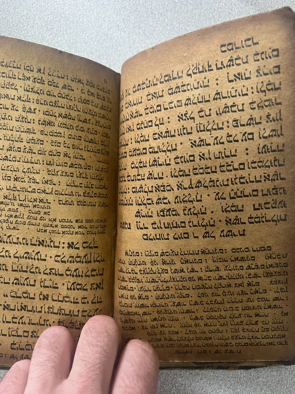 An open hand-written book in East Syriac script, held open by one hand, showing a large formal text block above a smaller denser one